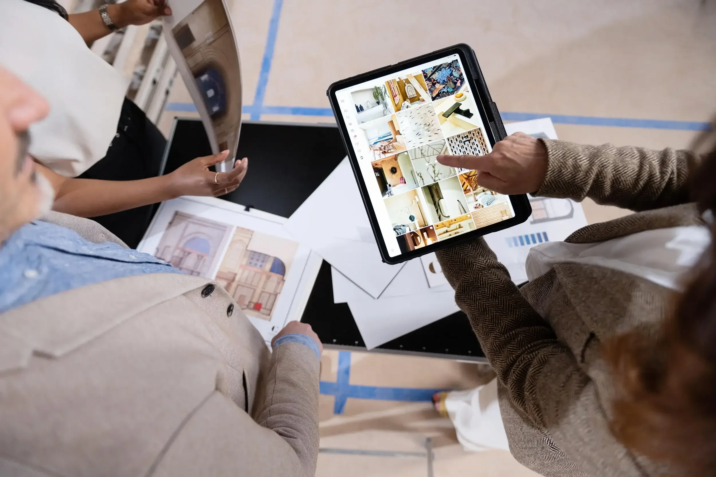 Two people look at digital devices displaying images of interior decor, surrounded by design plans and color swatches on a table.