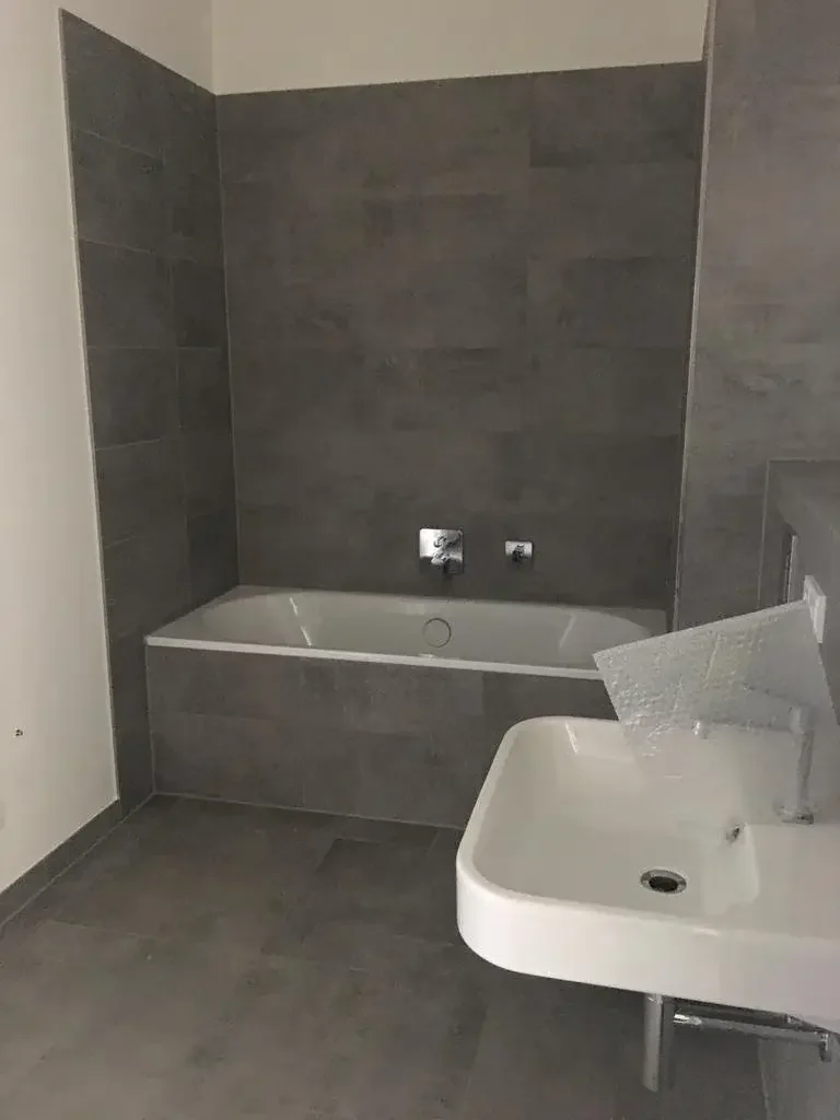 A bathroom with a bathtub on the back wall and a wall-mounted sink in the foreground, all with gray tile finishes.
