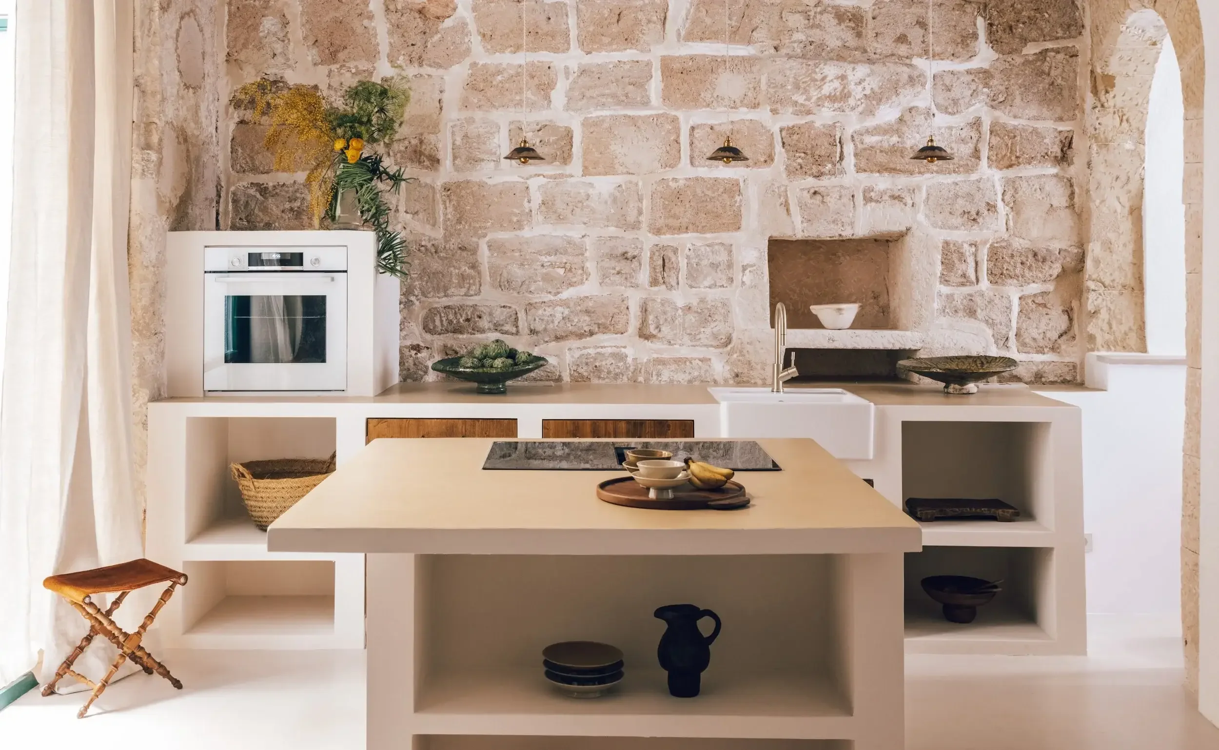 A kitchen with a rustic stone wall, white cabinetry, a built-in oven, and a beige countertop. Decor includes a vase with yellow flowers and greenery, a bowl of green artichokes, a serving tray with bowls and bananas, and various decorative pottery on