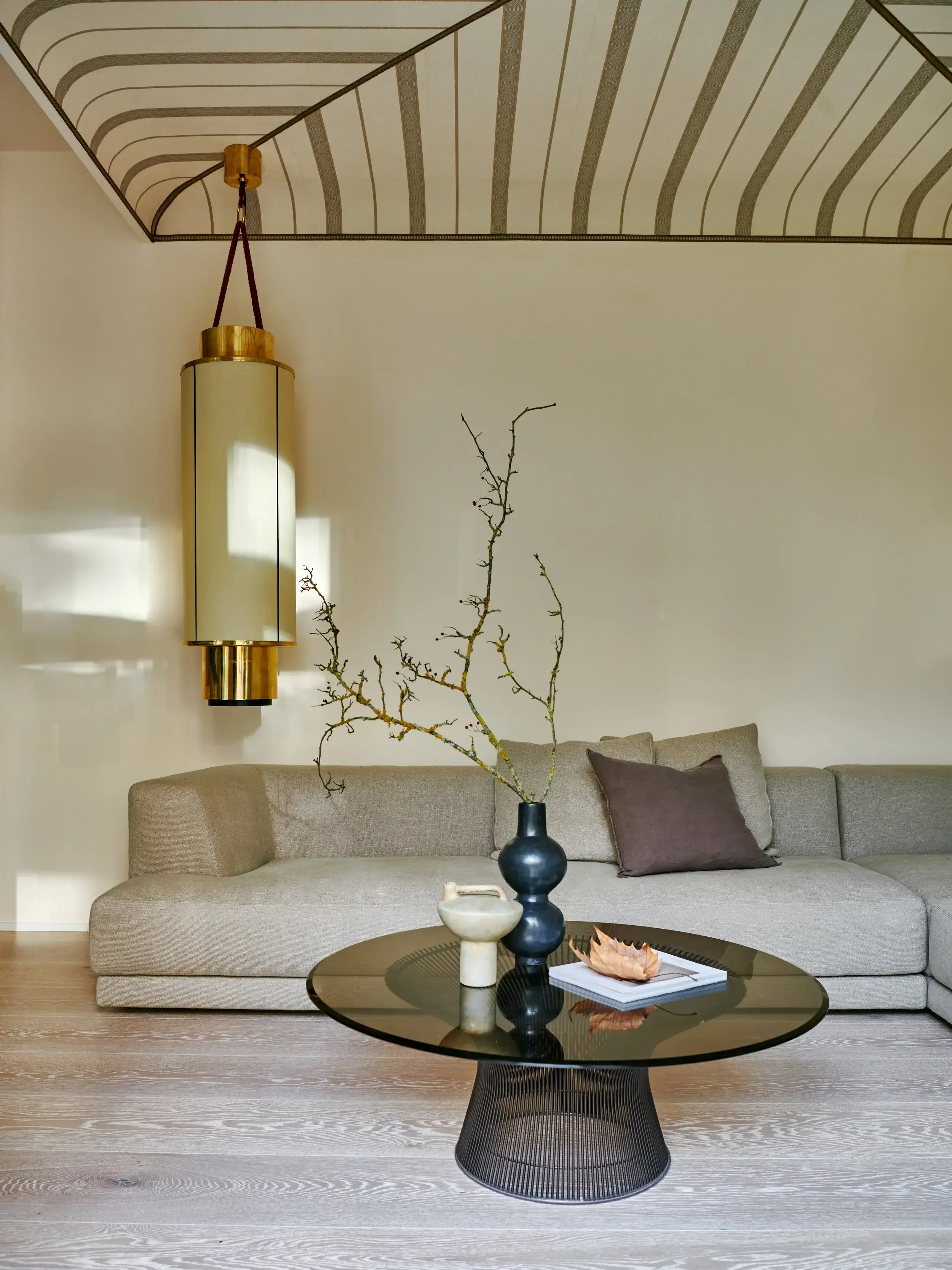 Modern living room with beige sofa, gray pillows, black round coffee table with decorative vases and a dried leaf, minimalist decor, and a gold wall sconce with cylindrical shade. Light wood floor and white wall.