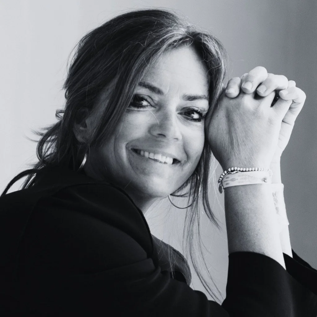 Schwarz-Weiß-Foto einer lächelnden Frau, die ihre Hände gefaltet an die Stirn gelegt hat. Sie hat schulterlanges Haar, trägt Creolen und ein Perlenarmband sowie ein dunkles Oberteil.