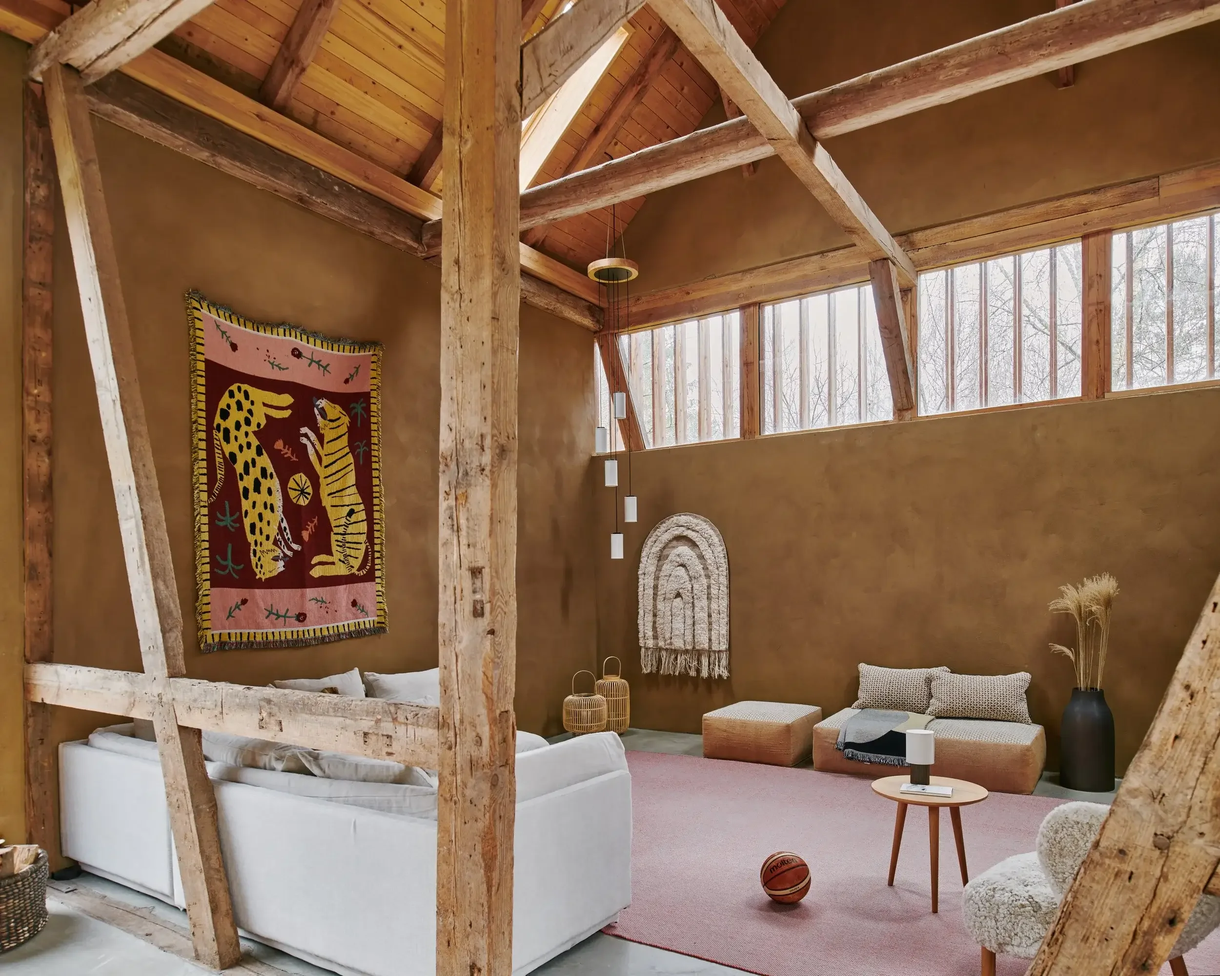 A cozy living room with a natural wood ceiling and walls, featuring a large white sofa, a small wooden round side table with black lamps, woven baskets, and a colorful tapestry with a leopard design hanging on the wall. There are large windows near t