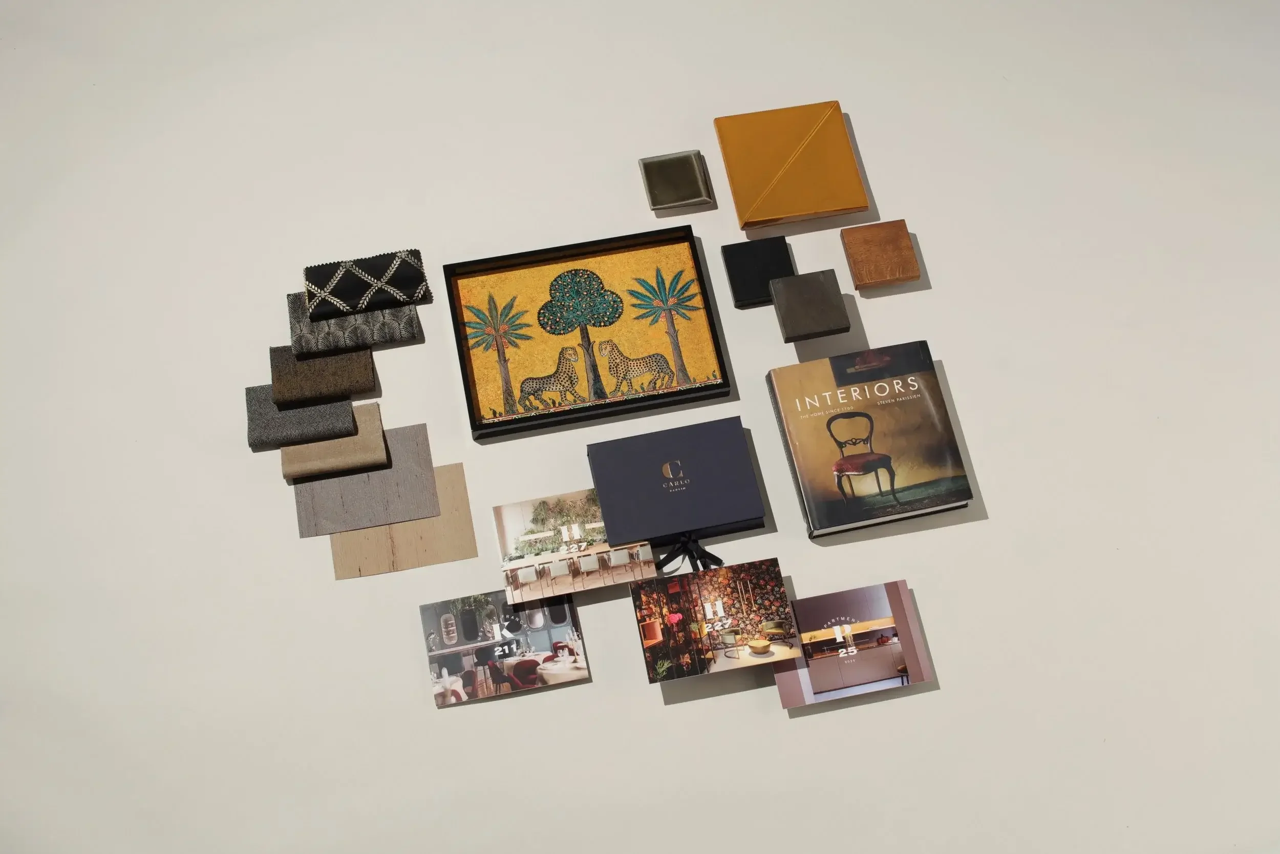 A flat lay of interior design magazines, fabric samples, and a framed artwork depicting two leopards under trees, arranged on a white surface.