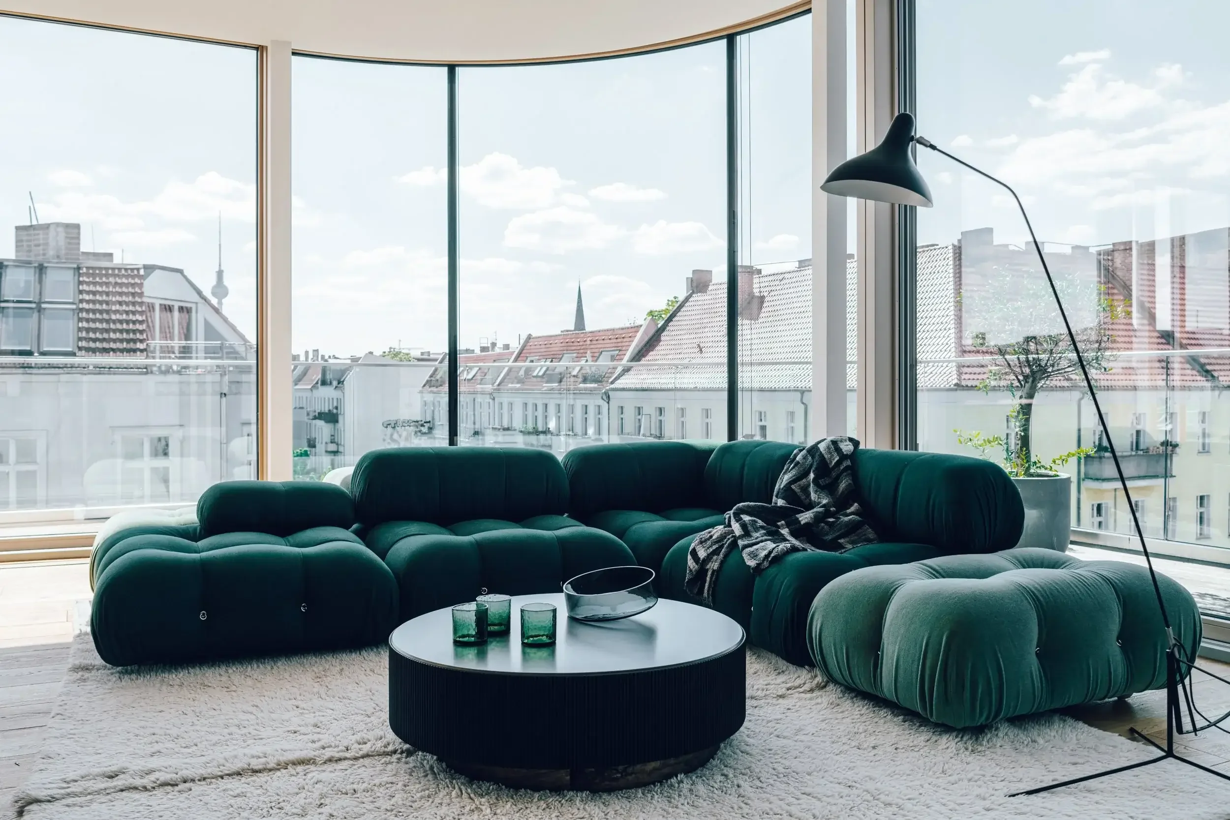 Living room with large floor-to-ceiling windows showing city buildings and sky, green velvet sectional sofa, black round coffee table with glasses, white shag rug, and a black floor lamp.