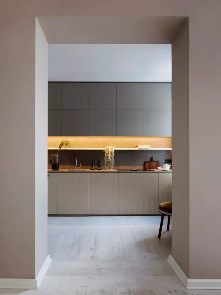 Modern kitchen with beige cabinets, dark backsplash, and open shelf with books, a plant, and a hanging plant. Counter has three bottles, a cutting board, and teapot set. Seen through doorway with a chair partially visible.