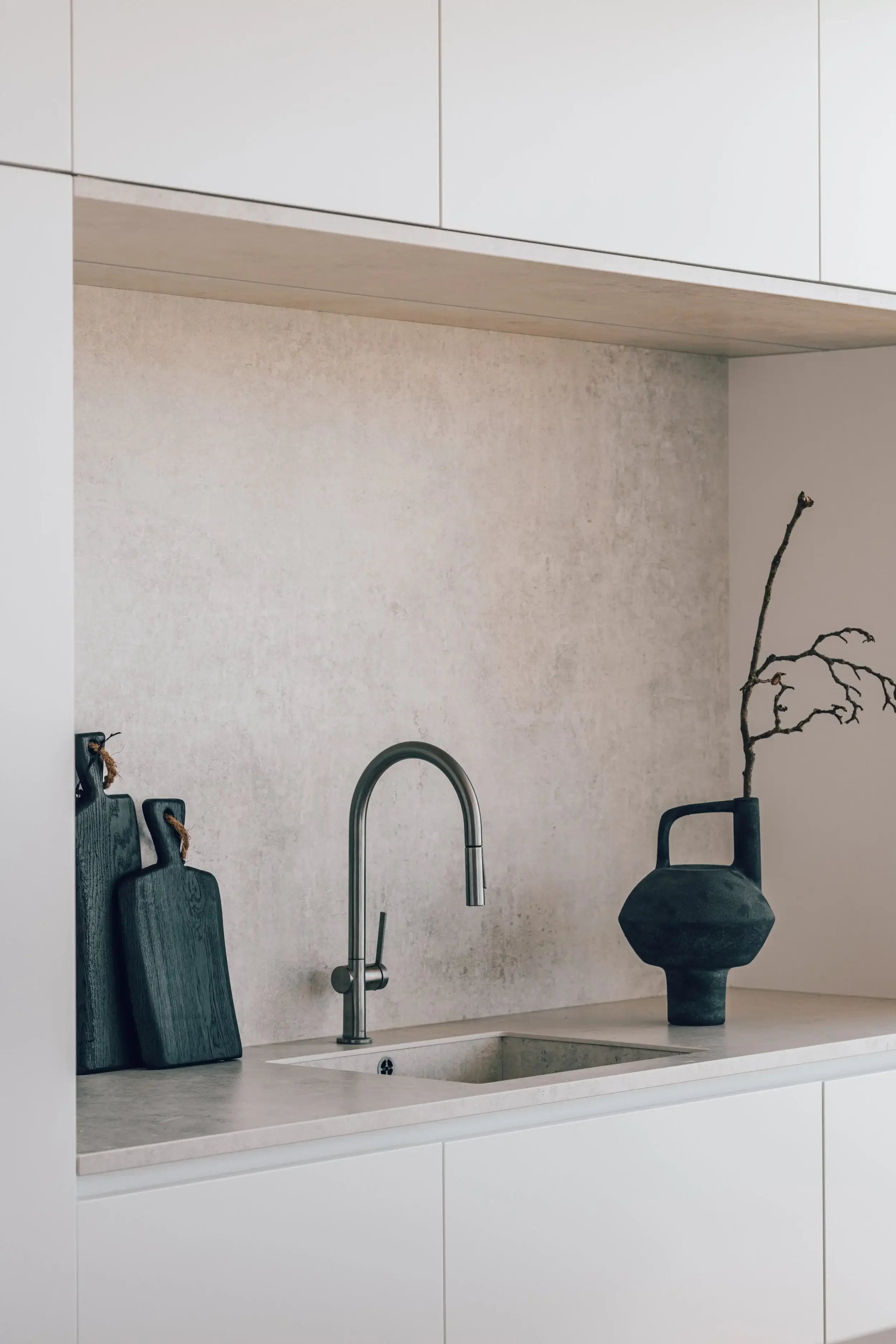 Minimalist kitchen countertop with a modern faucet, black cutting boards, and a black vase with a branch. Upper cabinetry and textured beige wall in the background.