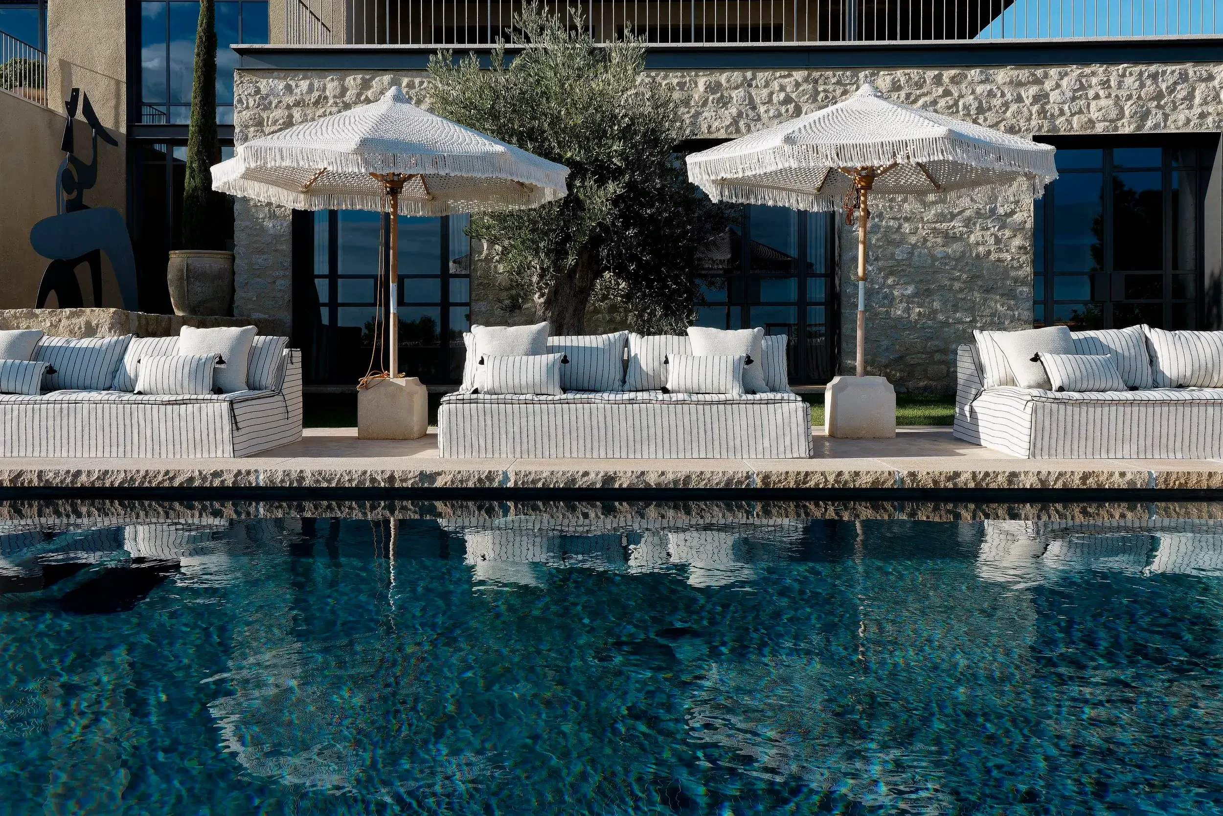 Outdoor seating area with striped cushioned sofas, two white fringed umbrellas, a pool in the foreground, and a modern building with large windows and stone walls in the background.