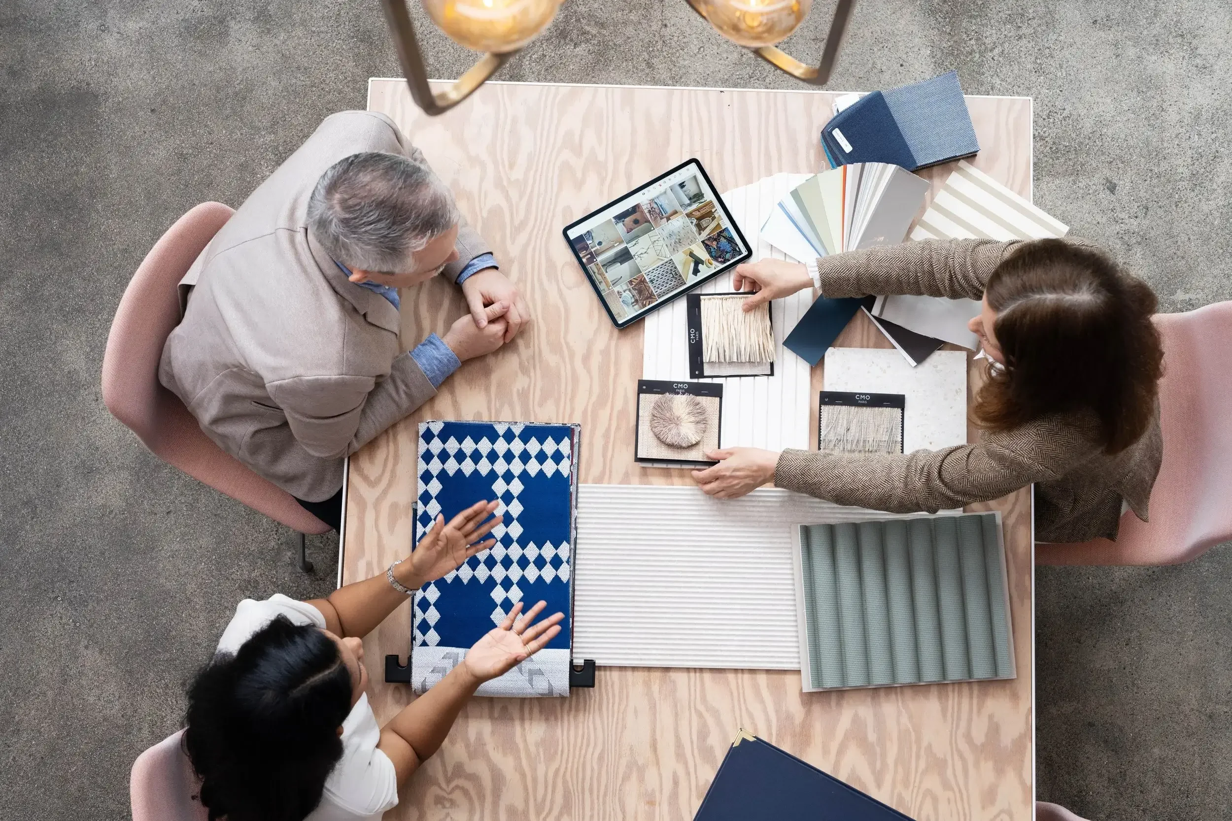 Three women and one child sitting around a wooden table discussing interior design material samples. The table has fabric swatches, paint color samples, a tablet, and sample books, with a concrete floor visible underneath.