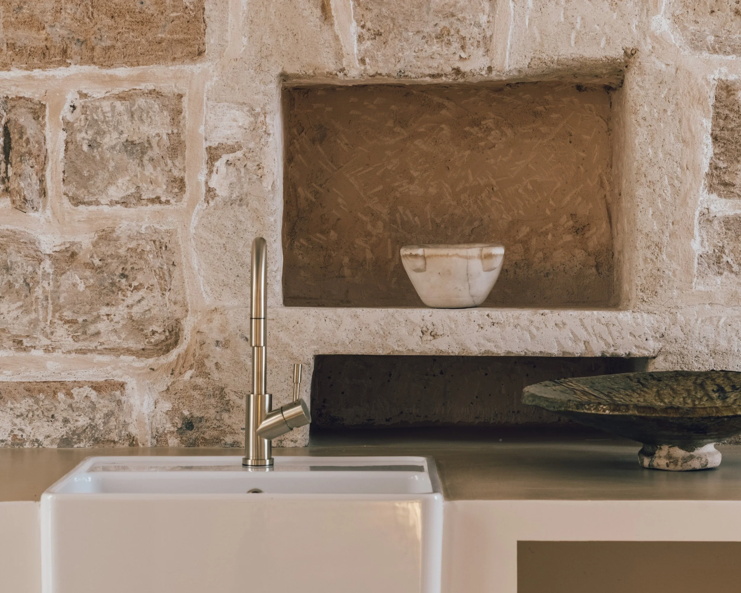 Kitchen countertop with a modern silver faucet, a white sink, a decorative bowl on the right, and a stone wall with a built-in niche holding a small ceramic bowl.