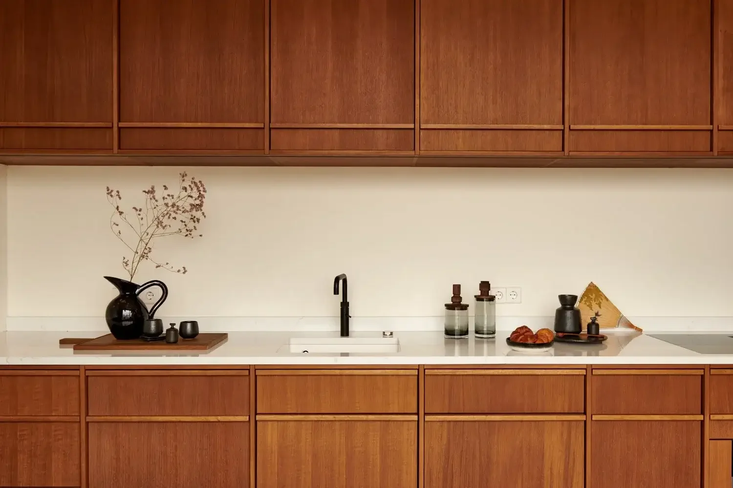Modern kitchen with wooden cabinets, white countertop, black sink faucet, black pitcher with dried branches, small black containers, and decorative items including glass bottles, a plate with bread, and a partially visible golden fan.
