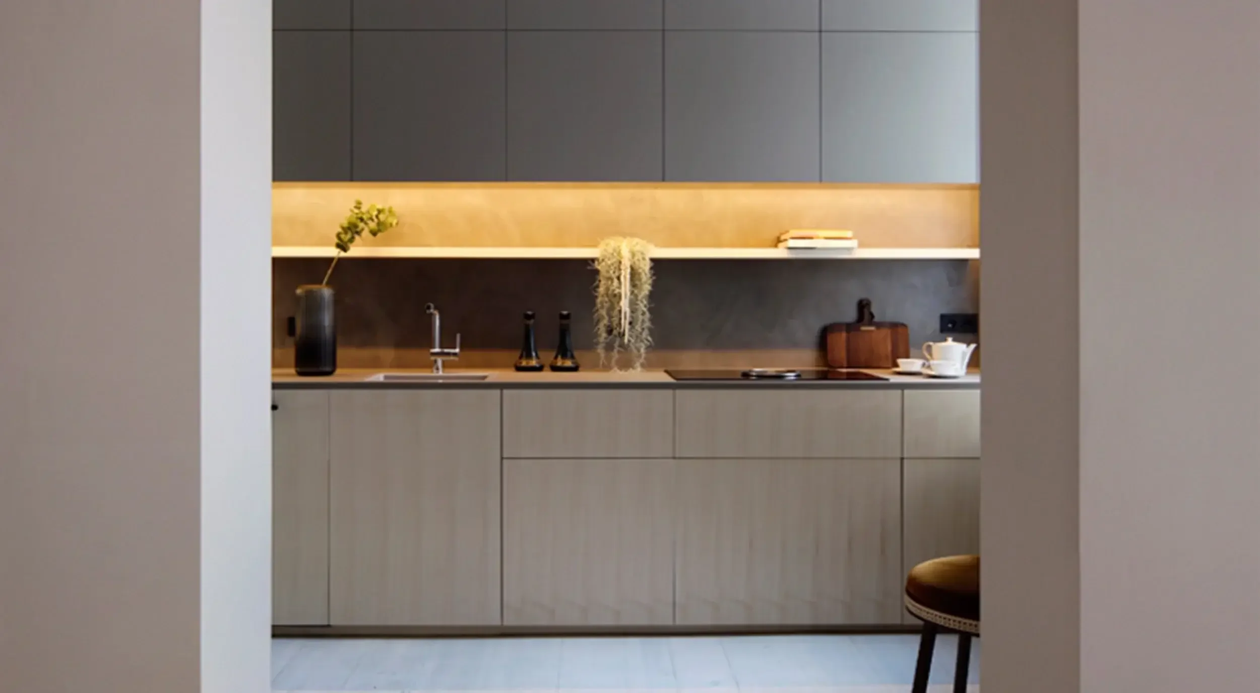 Modern kitchen with beige cabinets, dark backsplash, and open shelf with books, a plant, and a hanging plant. Counter has three bottles, a cutting board, and teapot set. Seen through doorway with a chair partially visible.
