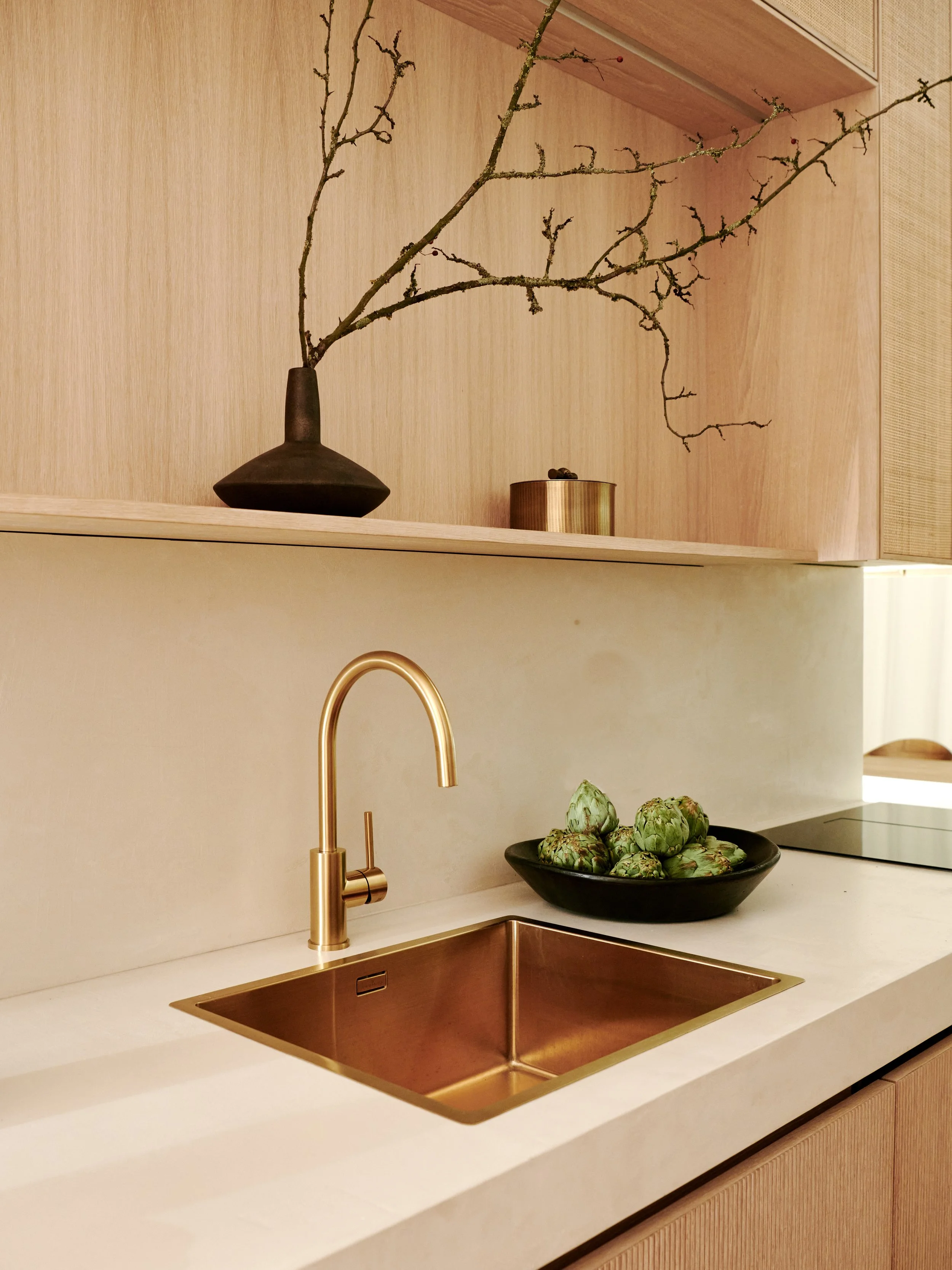 Modern kitchen with a white countertop, a gold sink, and a gold faucet. A black bowl with green artichokes on the counter. Wooden upper cabinet with a black vase containing branches and a gold container.