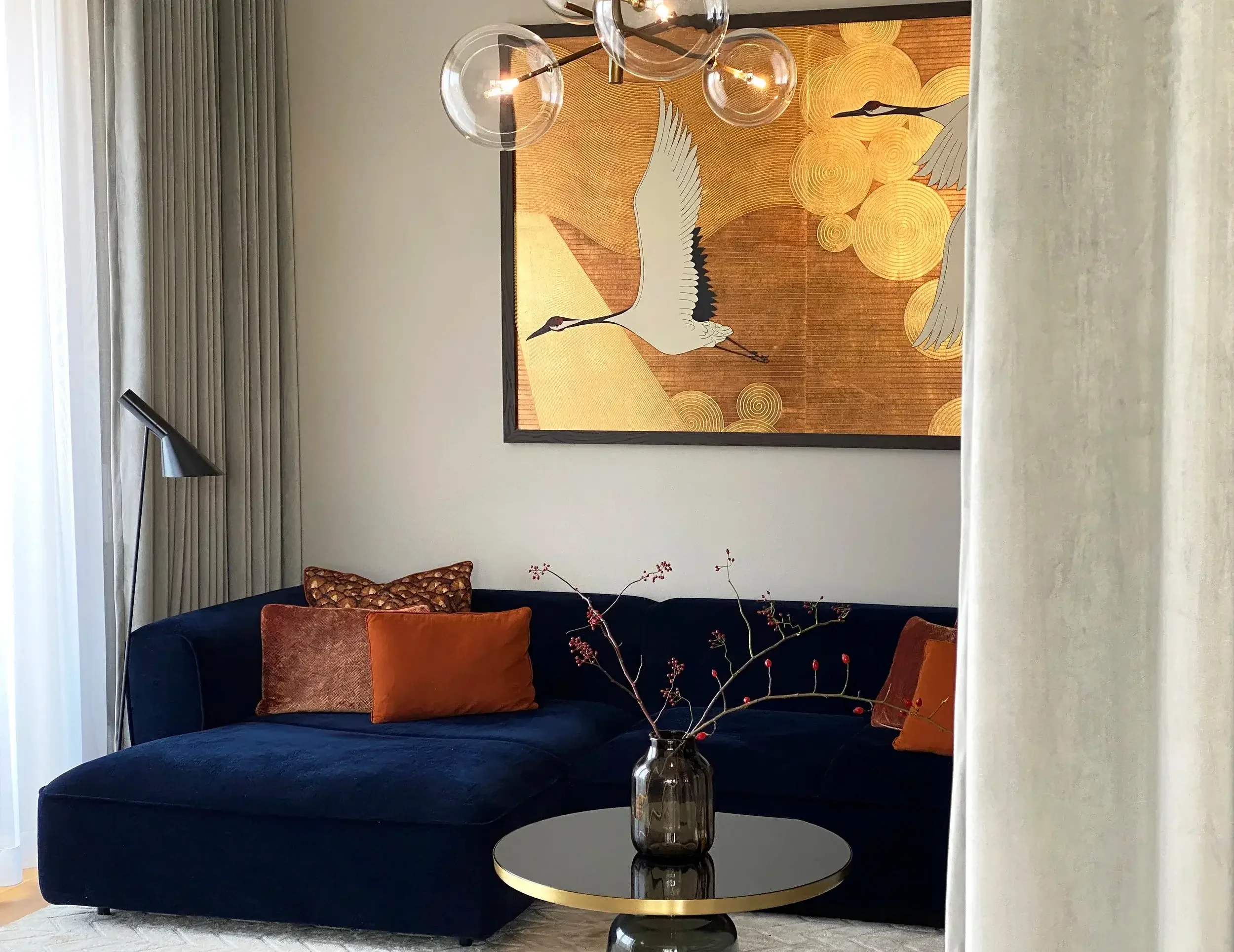 A living room corner featuring a dark blue velvet sofa with multiple pillows in shades of brown and orange, a round black coffee table with a decorative vase and branches, a large painting of cranes and gold circles on the wall, a modern black floor lamp, and hanging glass globe pendant lights.