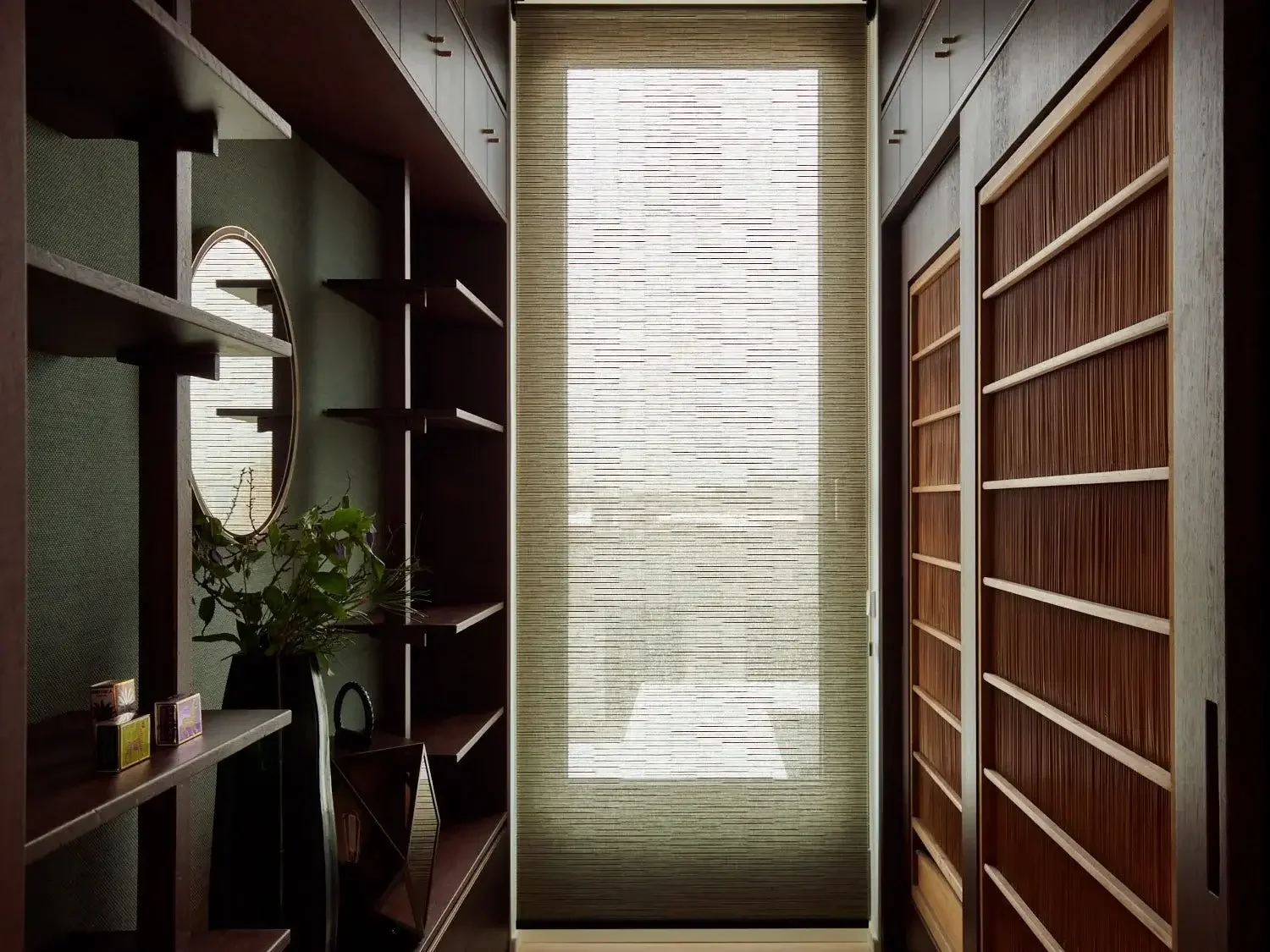 A narrow interior space with wooden shelving on the right, a green wall with a round mirror and small decorative items on the left, and a large window with a woven shade at the end of the corridor.