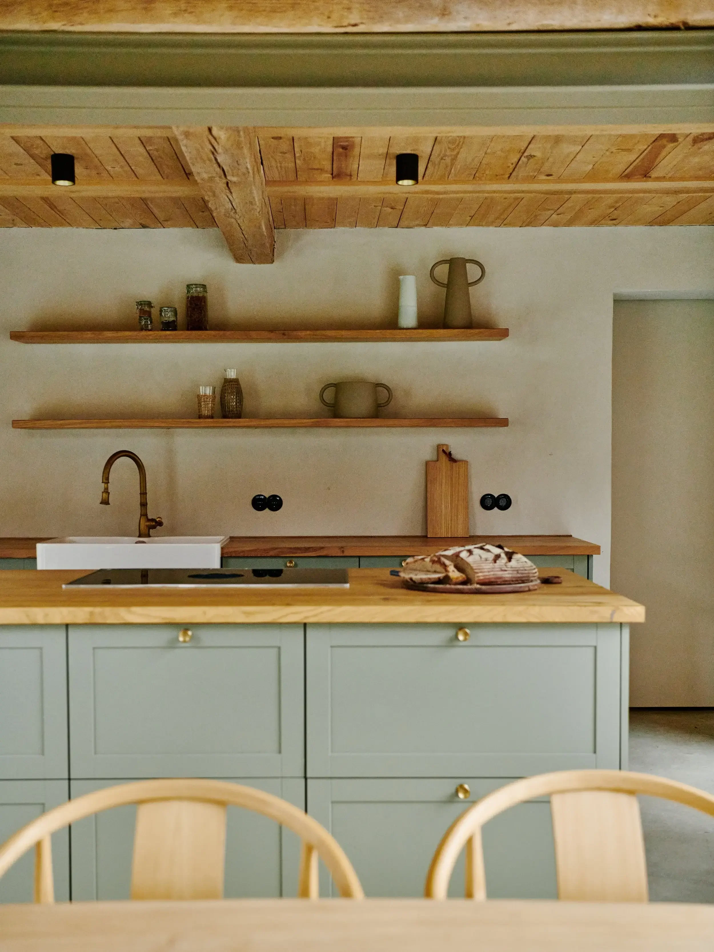 A modern kitchen with a green island cabinet, wooden countertop, and open wooden shelves holding jars and vases. There is a white farmhouse sink, a stovetop, and a loaf of bread on the counter. The ceiling is wooden with black recessed lights, and part of a wooden dining table with chairs is visible in the foreground.