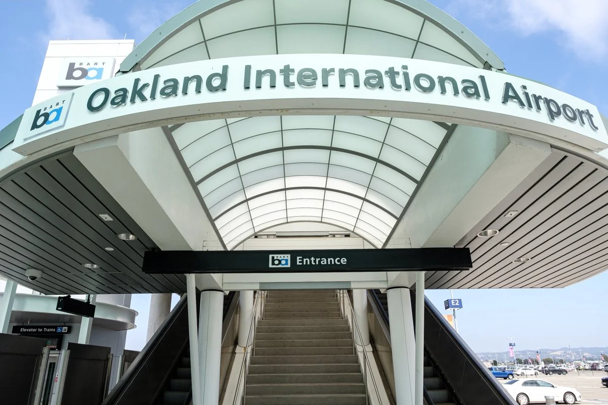 Entrance to Oakland International Airport with a curved glass canopy and an escalator leading up to the terminal. The sign reads 'Oakland International Airport' and 'Entrance'.