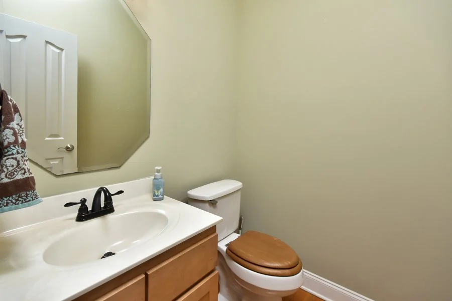 A small bathroom with a white sink, a wooden vanity, a mirror, a toilet with a brown seat cover, a soap dispenser, and a towel hanging on a hook.