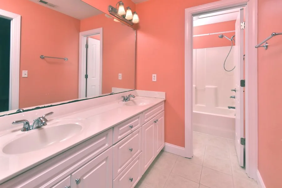 A bathroom with pink walls, a large mirror above a double sink vanity, and a shower/tub combo with a showerhead and soap dish.