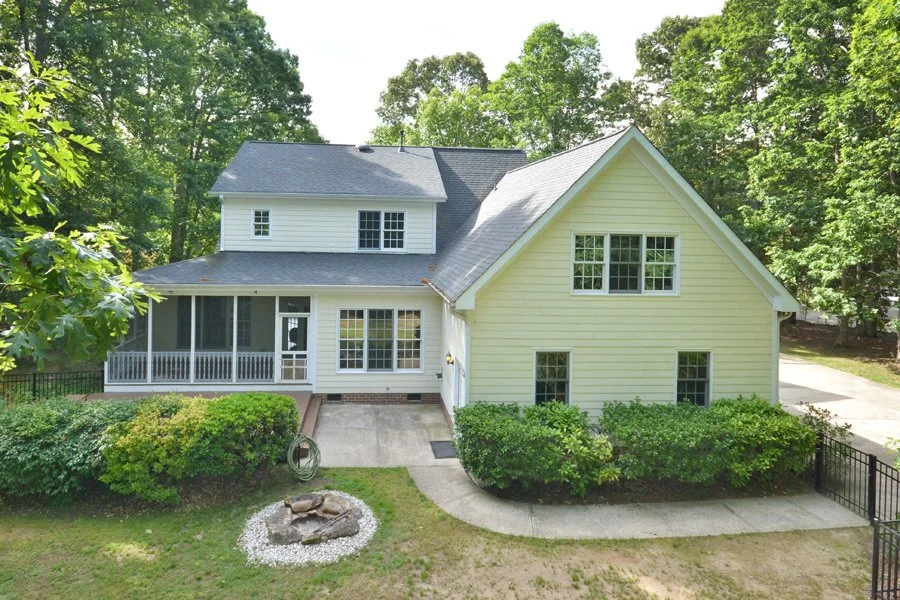 A two-story yellow house with a gray roof, surrounded by green trees and bushes, with a porch and a concrete pathway.