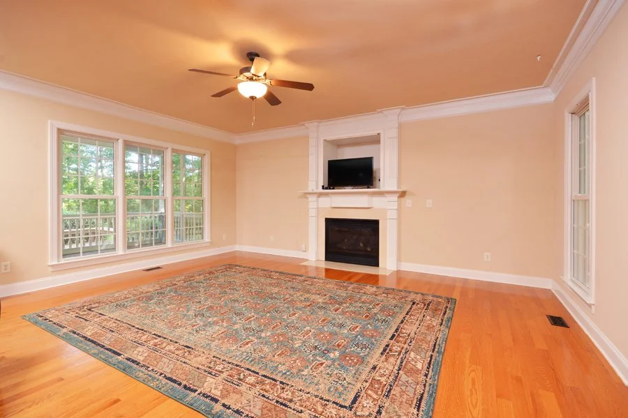 Empty living room with large windows, hardwood floors, a ceiling fan, a fireplace with a TV above, and a decorative area rug.