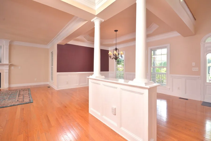 Interior of a spacious living room with hardwood floors, beige walls, white trim, a purple accent wall, and large windows. There is a chandelier hanging from the ceiling, and the room features decorative columns.