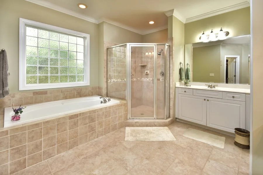 Bathroom with tiled bathtub, walk-in shower with glass door, large mirror, and white vanity with sink, green towels, window, and beige decor.