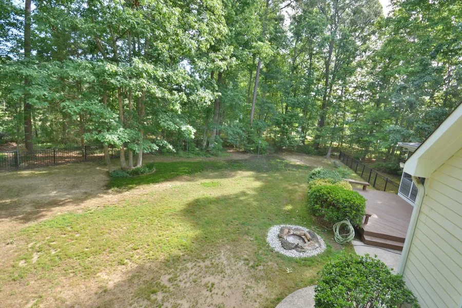 Backyard with grassy lawn, trees, bushes, a small fire pit, a patio with stairs, and a black fence in a suburban setting.