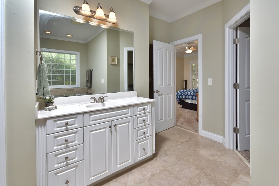 Bathroom vanity with a large mirror, sink, white cabinets, and a three-light fixture, with a towel hanging on the towel rack, a window, and a view into a bedroom with a bed and ceiling light.