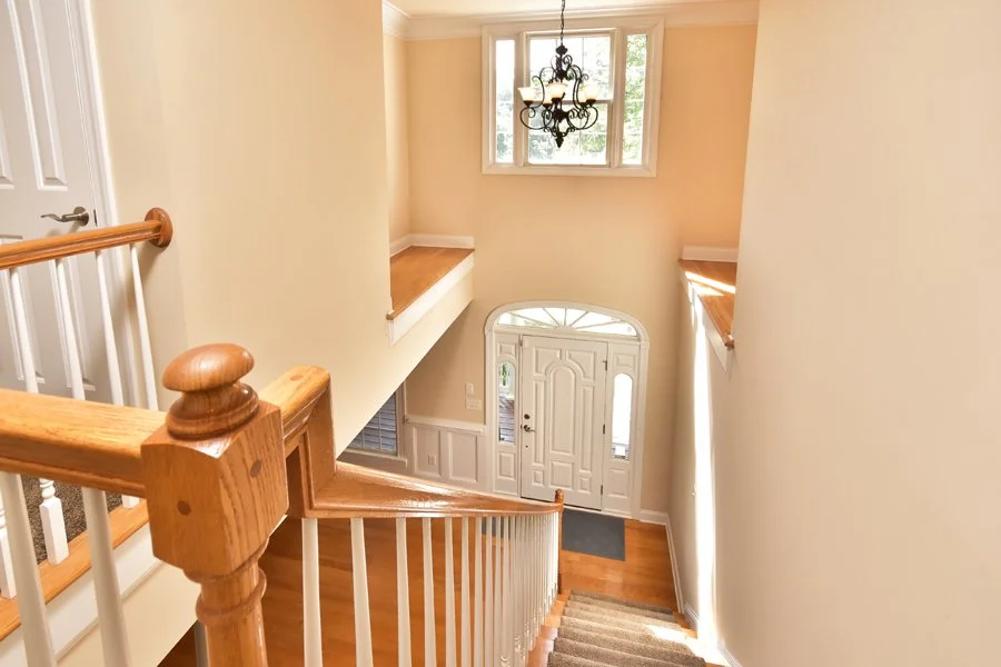 View of a spacious foyer from the top of a staircase, featuring a white front door with a decorative glass window, a chandelier hanging from the ceiling, and windows allowing natural light to illuminate the area.