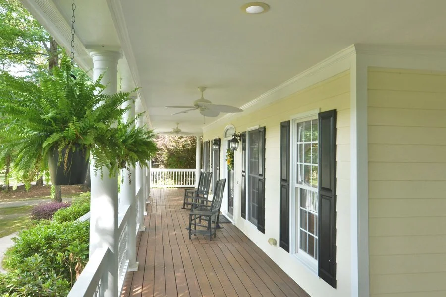 Front porch of a house with wooden flooring, white railing, hanging fern plant, yellow exterior wall with black shutters, two rocking chairs, ceiling fans, and a view of green trees and garden.