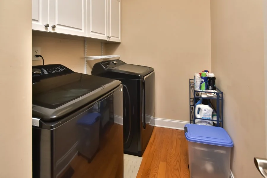 A laundry room with a top-loading washing machine and a front-loading dryer, white cabinets above, a small metal shelf with laundry supplies, and a blue storage bin on the floor.