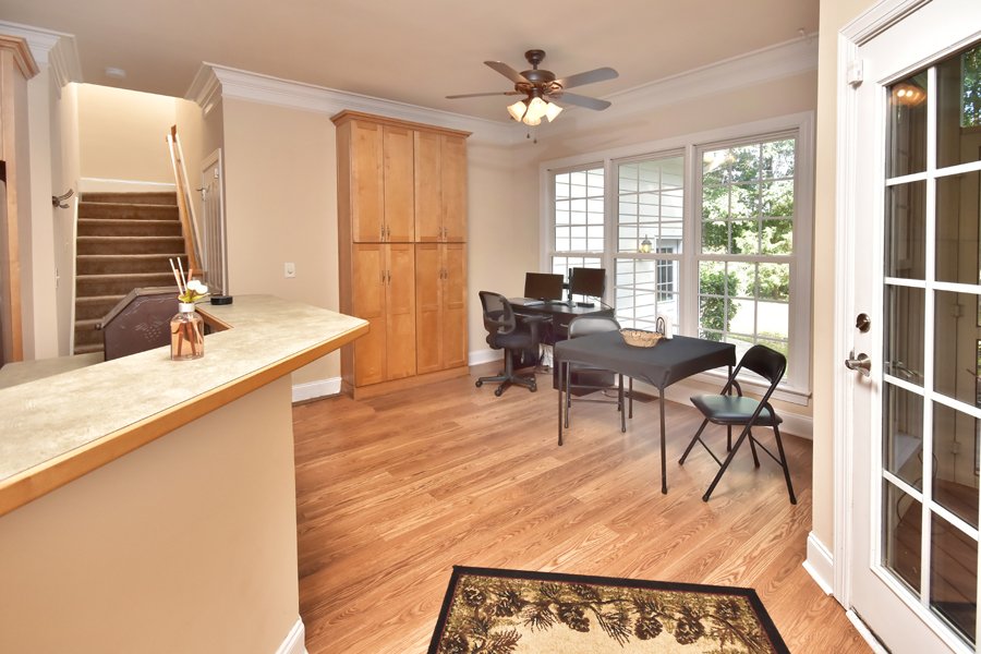 Bright dining area with large windows, wooden flooring, a ceiling fan, a wooden cabinet, a table with chairs, a black folding chair, a small black table, and a glass door leading outside.