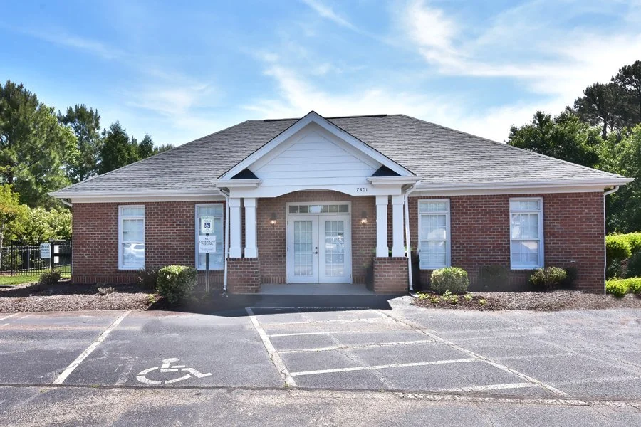 A single-story brick building with a white porch and a handicap parking space in front, surrounded by greenery under a partly cloudy sky.