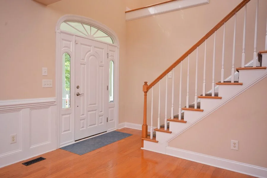 Interior view of a house entryway with a white front door, two narrow side windows, hardwood flooring, and a staircase with white spindles and a wooden handrail.
