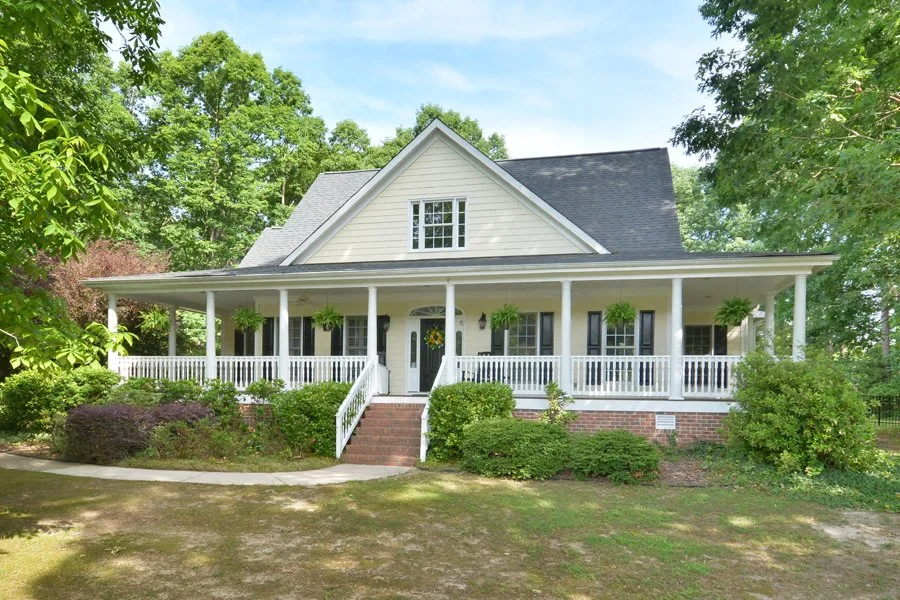 A large two-story house with a front porch, white walls, a dark shingle roof, and surrounded by trees and greenery.