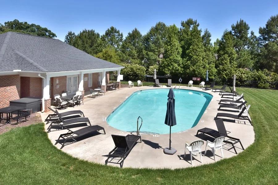 Outdoor swimming pool surrounded by lounge chairs and patio furniture, with a small building and lush green trees in the background under a blue sky.