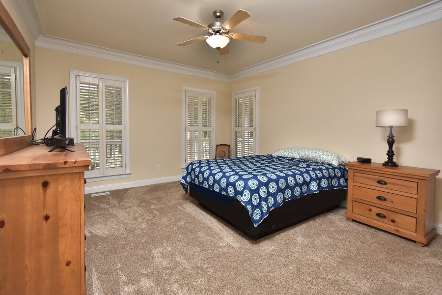 Spacious bedroom with a bed covered in a blue and white patterned quilt, wooden nightstand with a lamp, wooden dresser with a television, and multiple windows with white shutters.
