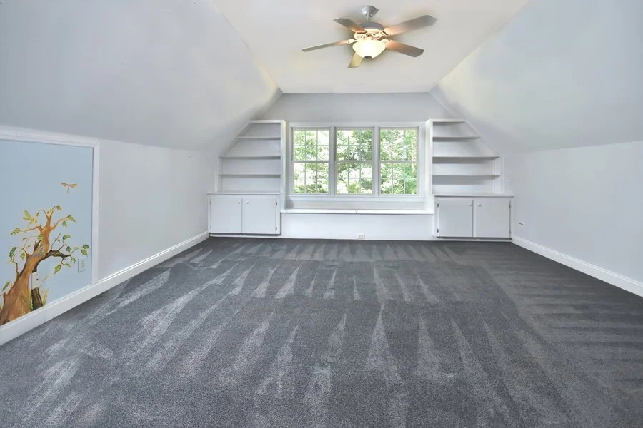 Empty attic bedroom with gray carpet, white walls, built-in bookshelves, ceiling fan, and a large window overlooking trees.