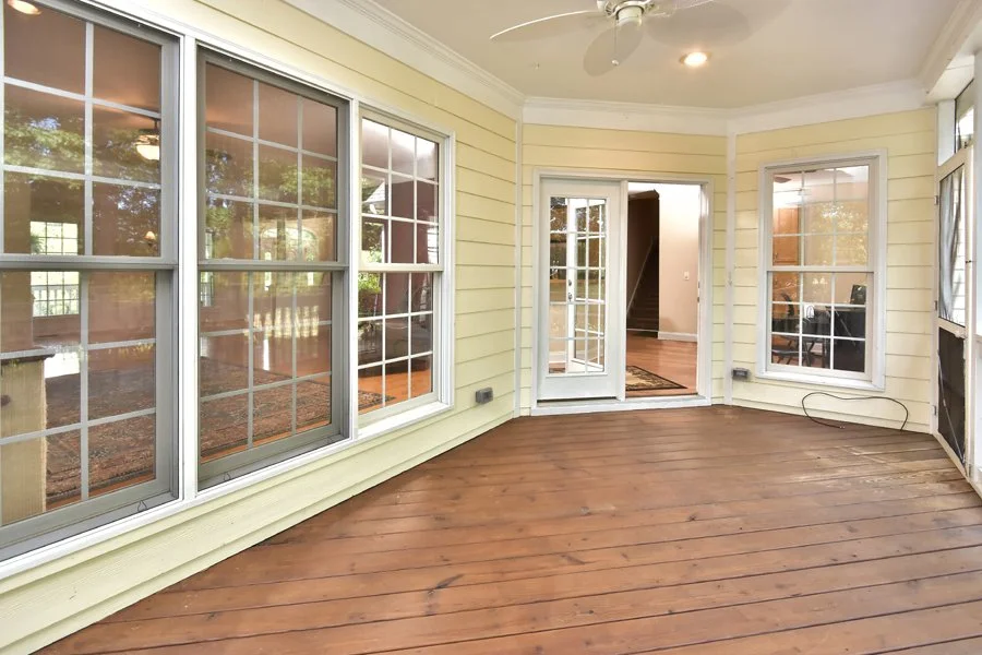 A screened-in porch with wooden flooring, white framing, large windows, a glass door leading to the interior of a house, and a ceiling fan.