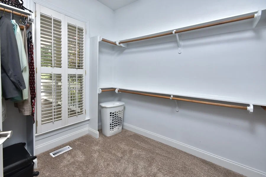Empty walk-in closet with white walls, beige carpeting, empty wooden closet rods, a laundry basket, and a large window with shutters.