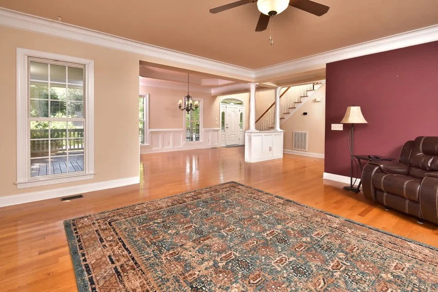 Living room with hardwood floors, a large area rug, beige walls, a maroon accent wall, a brown leather recliner, a floor lamp, a ceiling fan, and windows showing a deck and greenery outside.