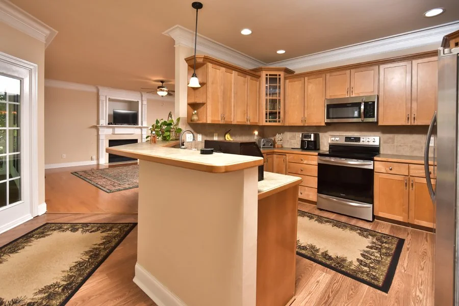Kitchen with wooden cabinets, a stainless steel refrigerator, oven, microwave, and small appliances on the countertop. An island with a sink and pendant light, cushioned chairs, and a view into the living room with a fireplace and television.