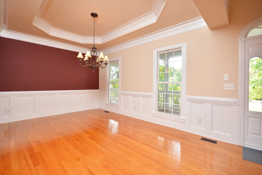 Empty dining room with hardwood floors, beige and maroon walls, chandelier lighting, white crown molding, and three large windows with view of green trees outside.