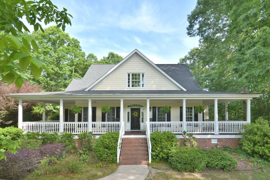 Front view of a white house with a porch surrounded by lush greenery and trees, a waving American flag on the left, and a white sign with red and blue lettering indicating an open house event for Memorial Day themed on May 24, 2026, from 1 PM to 3 PM.