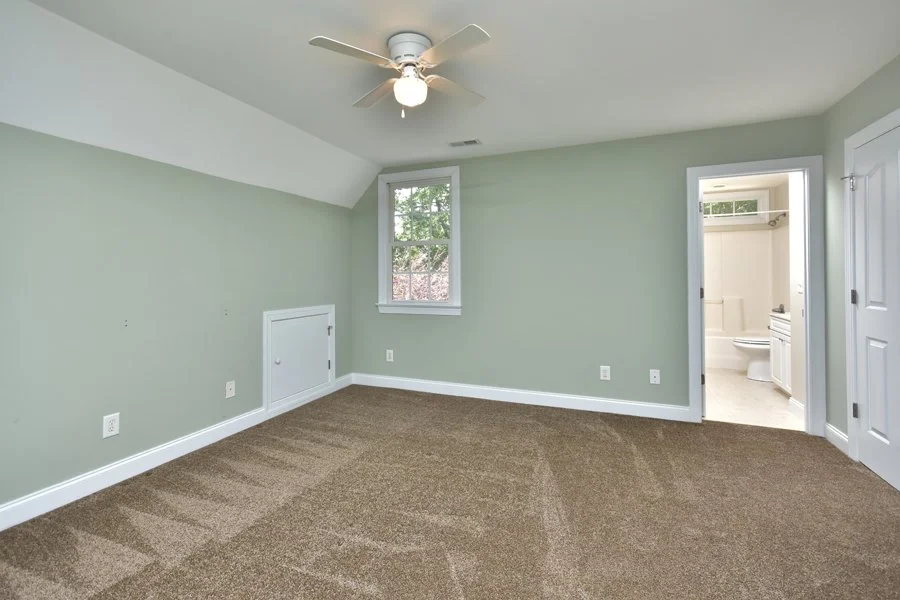 Empty bedroom with green walls, ceiling fan, brown carpet, and an adjacent bathroom.