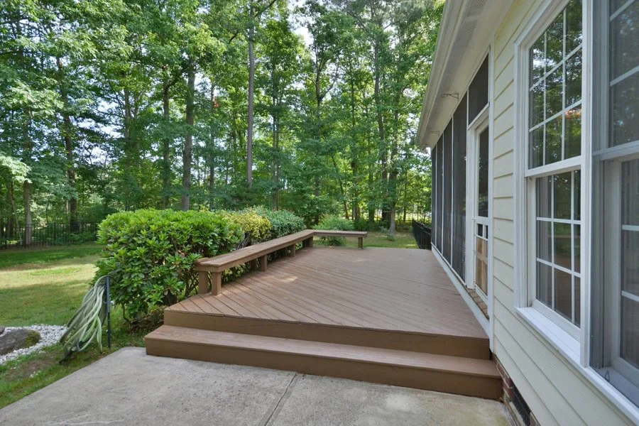 Wooden deck attached to a house, with steps leading to a concrete patio, surrounded by greenery and trees.