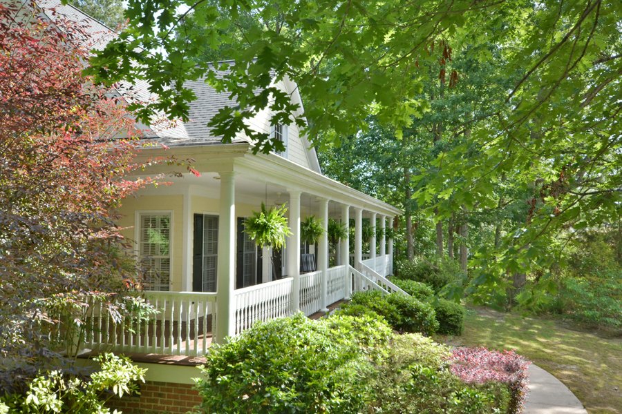 White house with a porch, surrounded by lush green trees and shrubs, with hanging plants and a curved pathway.
