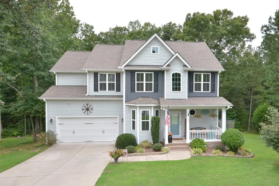 Front view of a two-story suburban house with a gray and blue exterior, white trim, a two-car garage, and a small front porch adorned with plants and an American flag.