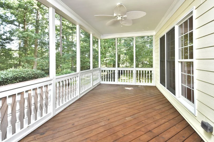 A spacious screened-in porch with wooden flooring, white railing, and ceiling fan, overlooking a lush green wooded area.