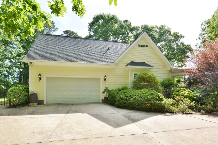 Yellow house with a two-car garage, surrounded by green bushes and trees, with a concrete driveway.